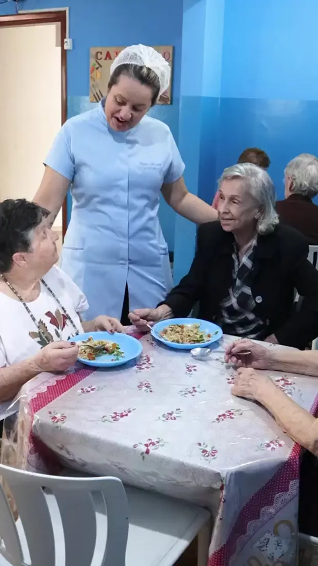 Cuidadora serve a refeição a duas senhoras sentadas à mesa